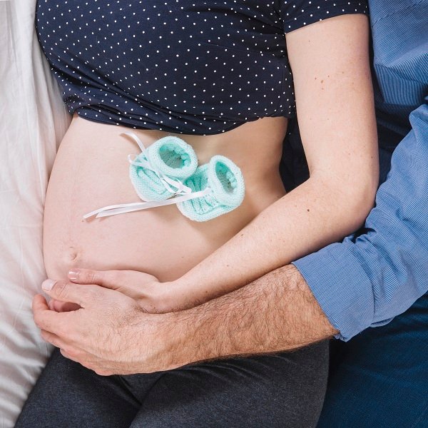A close-up photo of a pregnant woman’s belly with baby shoes placed on it, while her partner lovingly rests his hand on her stomach.